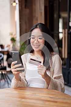 A happy Asian woman sits at an outdoor table of a cafe, holding her credit card and smartphone