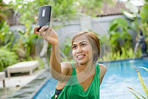happy Asian woman taking selfie picture for social media internet app on mobile phone camera relaxed at resort swimming pool
