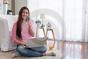 Happy asian woman talk on mobile and  typing laptop computer at home.female freelancer sitting on carpet on floor work at home in