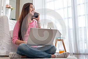 Happy asian woman talk on mobile and  typing laptop computer at home.female freelancer sitting on carpet on floor work at home in