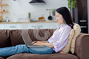 Happy Asian woman using laptop on sitting of cozy sofa at home chatting on social network