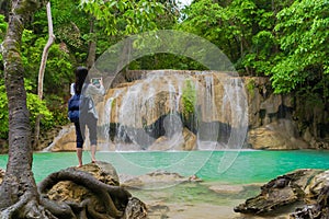 Happy Asian woman using mobile phone to take a photo by camera on social media at Erawan waterfall in tropical forest with trees