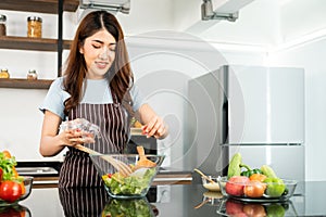 Happy Asian woman wearing apron, adding cherry tomato while tossing the vegetarian salad with wooden spatulas.