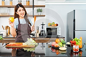 Happy Asian woman wearing apron, holding chili pepper and wooden spatula. Preparing a healthy salad with fresh vegetables.