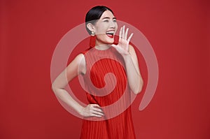 Happy Asian woman wearing red dress with open mouths raising hands shouting announcement isolated on red copy space background