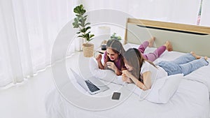 Happy Asian women couple laying in bed using laptop for online shopping together in the morning with a smile and laugh.