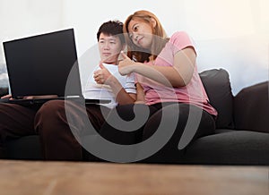 Happy Asian young couple sit relaxing on sofa watching laptop and show thumbs up sight at home