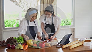 happy asian young daughter and senior mother cooking online class on tablet together making fresh vegetables food in kitchen at