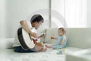 Happy Asian Young Father and Adorable little boy son sitting on bed playing acoustic guitar together at home