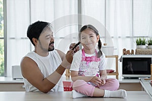 Happy Asian young father with beard making braid for his Caucasian little daughter while sitting on table at home kitchen