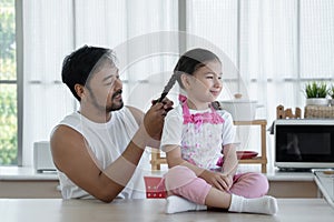 Happy Asian young father with beard making braid for his Caucasian little daughter while sitting on table at home kitchen