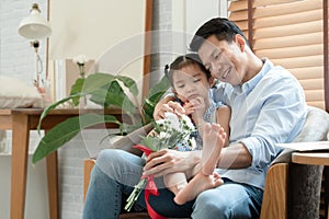 Happy Asian young father getting bunch of flowers congratulations on Fathers Day from little cute daughter sitting on his lap