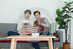 Happy Asian young gay couple wearing sweater holding joysticks playing game with excited face have fun together at living room