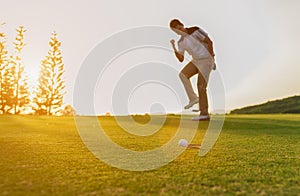 Happy asian young man golf player glad and showing gesture after putting golf ball down hole for birdie on golf green and outdoor.