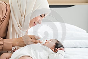Happy Asian young Muslim mother wear hijab holding milk bottle, feeding milk her cute newborn baby 1 month old, lying on bed at