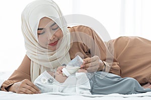 Happy Asian young Muslim mother wear hijab holding milk bottle, feeding milk her cute newborn baby 1 month old, lying on bed at