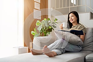 happy asian young woman sitting on sofa and reading book at home -people and leisure concept
