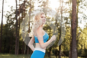 Happy athletic caucasian young woman blonde with cute smile jogging on background of trees and bright summer sunset in park.