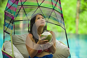 Happy and attractive Asian Chinese woman sitting at hanging hammock drinking coconut water relaxing at resort swimming pool