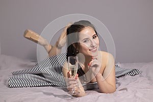 Happy attractive brunette woman in evening cocktail dress with a glass of champagne on a bed in grey bedroom. afterparty,