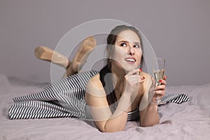 Happy attractive brunette woman in evening cocktail dress with a glass of champagne on a bed in grey bedroom. afterparty,