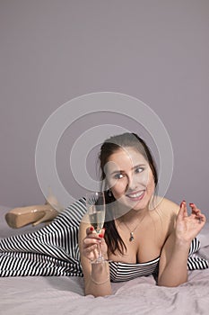 Happy attractive brunette woman in evening cocktail dress with a glass of champagne on a bed in grey bedroom. afterparty,