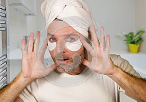 Happy and attractive camp gay man applying moisturizer eye patch facial product with head wrapped in towel looking playful