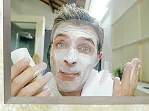 Happy and attractive Caucasian man at home looking at himself on bathroom mirror applying white facemask in male beauty and facial