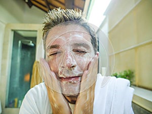 Happy and attractive Caucasian man smiling fresh at home bathroom washing his face with exfoliant soap looking at himself in toile