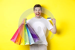 Happy attractive man holding shopping bags, showing credit card, standing over yellow background