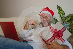 Happy and attractive man in Santa hat using credit card and laptop computer buying online Christmas presents holding gift box