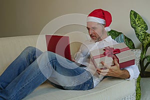 Happy and attractive man in Santa hat using credit card and laptop computer buying online Christmas presents holding gift box