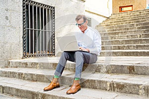 Happy attractive mature man working on laptop checking email sitting on stairs outdoors urban area
