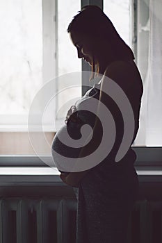 Happy Attractive pregnant woman standing near the window and holding her belly