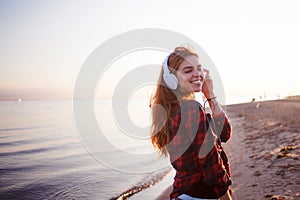 Happy attractive red-haired girl is enjoying music on walk. Beautiful young woman in shirt uses headphones, soft focus