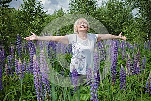 Happy attractive senior woman enjoys walk in purple lupines in blooming field, active in retirement