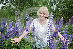 Happy attractive senior woman enjoys walk in purple lupines in blooming field, active in retirement