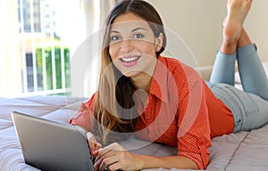 Happy attractive student girl using notebook lying on bed looking at camera. Homeschooling, e-learning, smart school concept