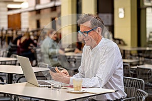 Happy attractive stylish mature business man working on laptop showing thumb up gesture outside cafe