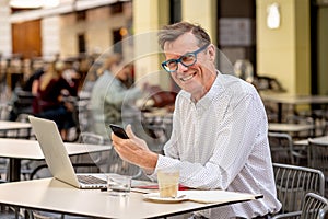 Happy attractive stylish mature man sending message on mobile while working on laptop outside cafe