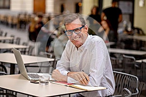 Happy attractive stylish mature man working on laptop while having coffee in outside coffee shop