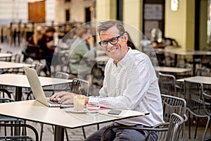 Happy attractive stylish mature man working on laptop while having coffee in outside coffee shop