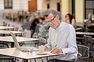 Happy attractive stylish mature man working on laptop while having coffee in outside coffee shop