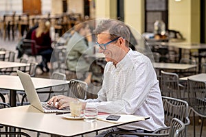 Happy attractive stylish mature man working on laptop while having coffee in outside coffee shop