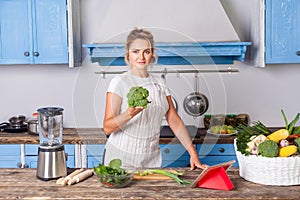 Happy attractive woman in apron holding broccoli and smiling at camera, cooking salad in modern kitchen