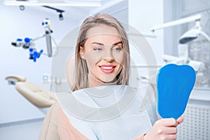 Happy attractive woman looking at her teeth in mirror. Satisfied patient of dental care or whitening for dental treatment