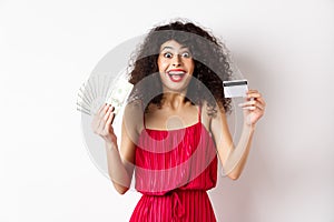 Happy attractive woman in red dress, screaming of joy and showing plastic credit card with money, winning prize