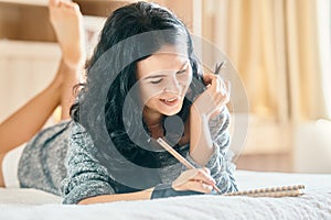 Happy attractive woman smiling and drawing with pencil in notebook while lying on the sofa