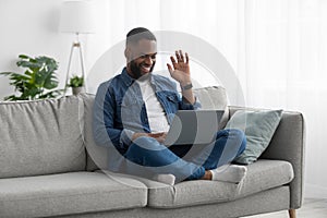 Happy attractive young african american male with beard sits on couch, greets, waves his hand to pc webcam