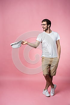 Happy attractive young man holding hat and asking for money over pink background.